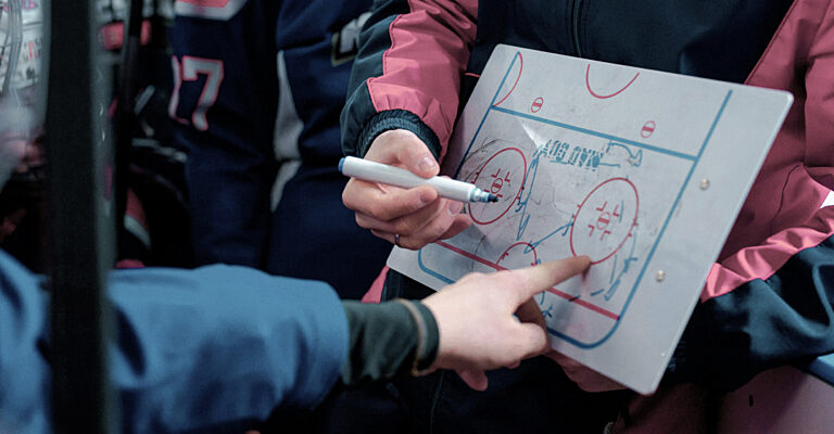 Coach holding an ice-hockey tactics board and drawing a play while a player points, at the rink.