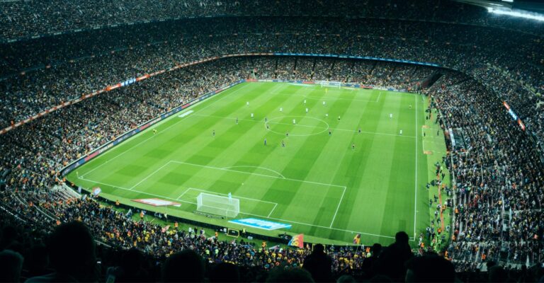 A packed football stadium at night with full stands and players on the floodlit pitch, capturing the emotional scale of live sport.
